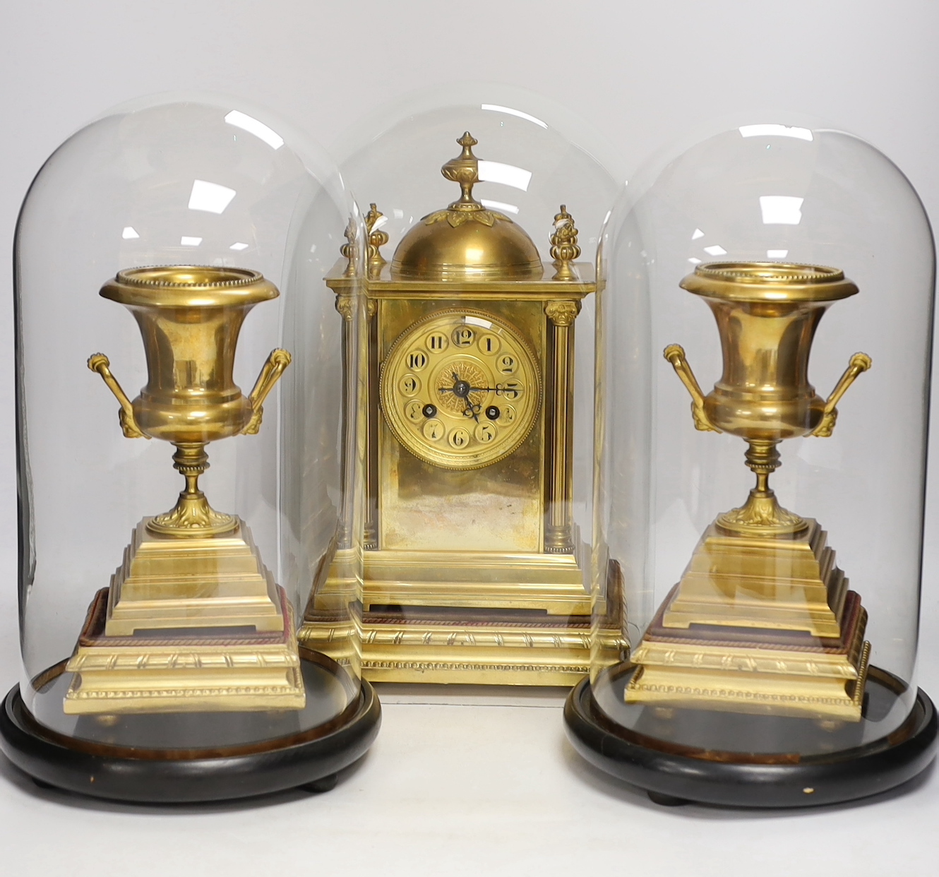 A gilt metal clock garniture, c.1900, with a pair of urns, all under glass domes, French movement by A D Mougin, striking on a gong, (missing base for clock dome), 36cm high